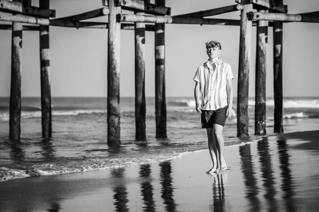 Black and white photo of a young man at his senior portrait session walking on the beach